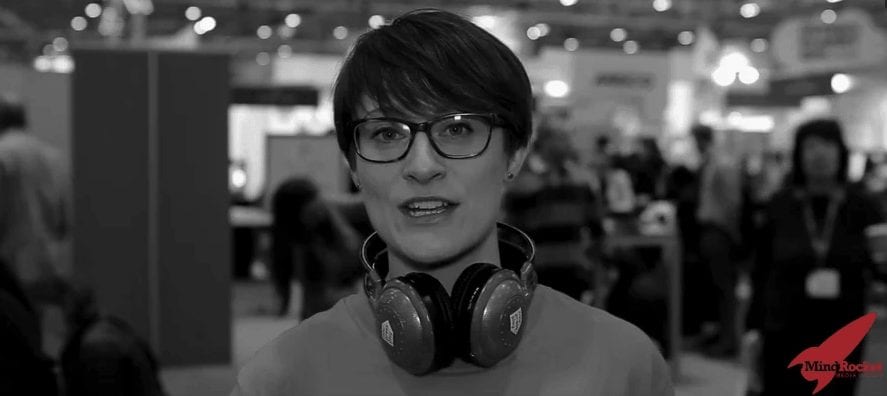 A person with short hair, glasses, and headphones around their neck is speaking at an indoor event or conference, with a crowd and booths blurred in the background.