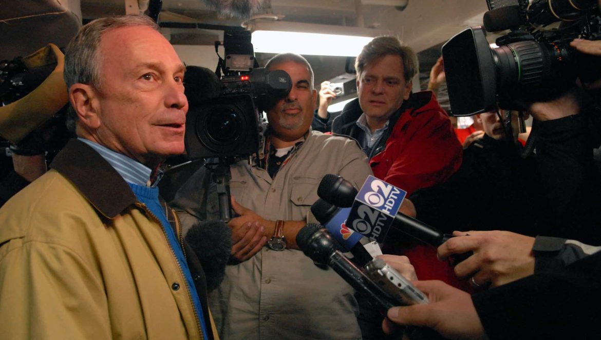 A man in a tan jacket speaks to a group of reporters holding microphones and cameras, surrounded by bright lights and recording equipment in an indoor setting.