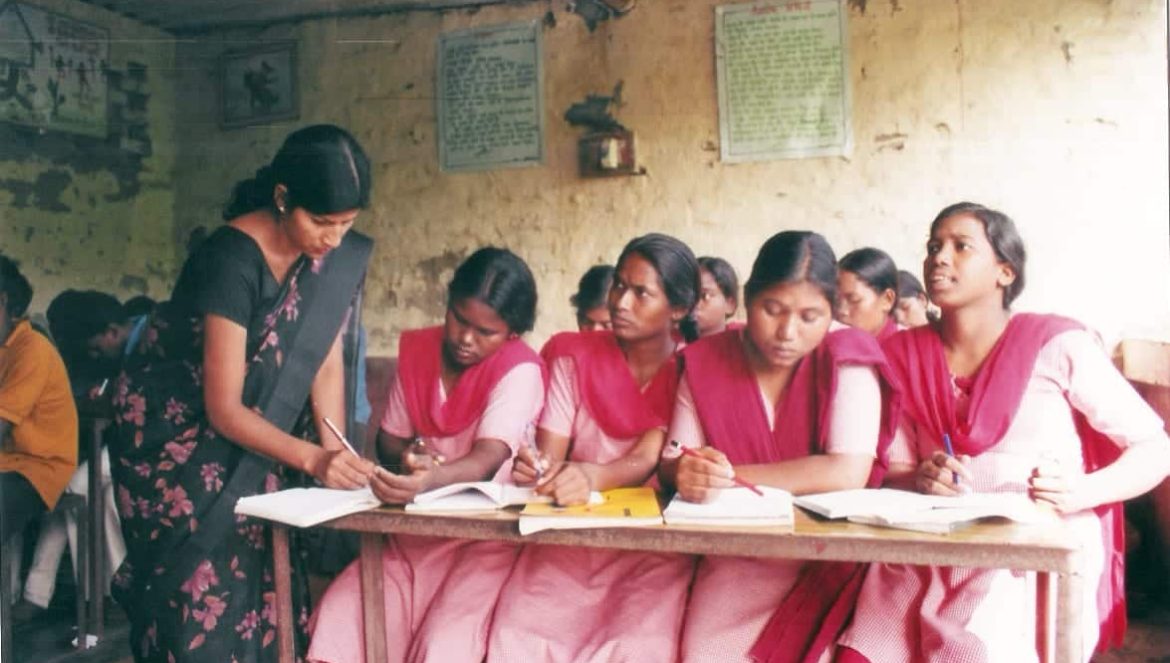 A teacher helps four female students in pink and red uniforms with their work in a classroom with worn walls and educational posters. The students sit at a wooden desk, focused on their notebooks.