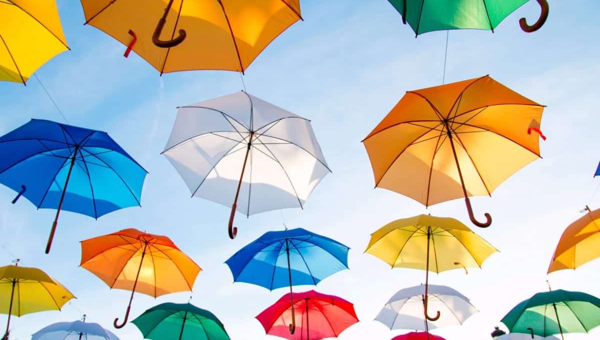 Colorful umbrellas, including yellow, blue, orange, green, and white, are suspended in the air against a clear blue sky, creating a vibrant and lively display.