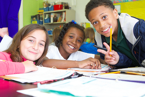 Three smiling children sit at a table with open notebooks and pencils, appearing happy and engaged in a classroom setting. Two girls and one boy face the camera, one making a peace sign with his fingers.