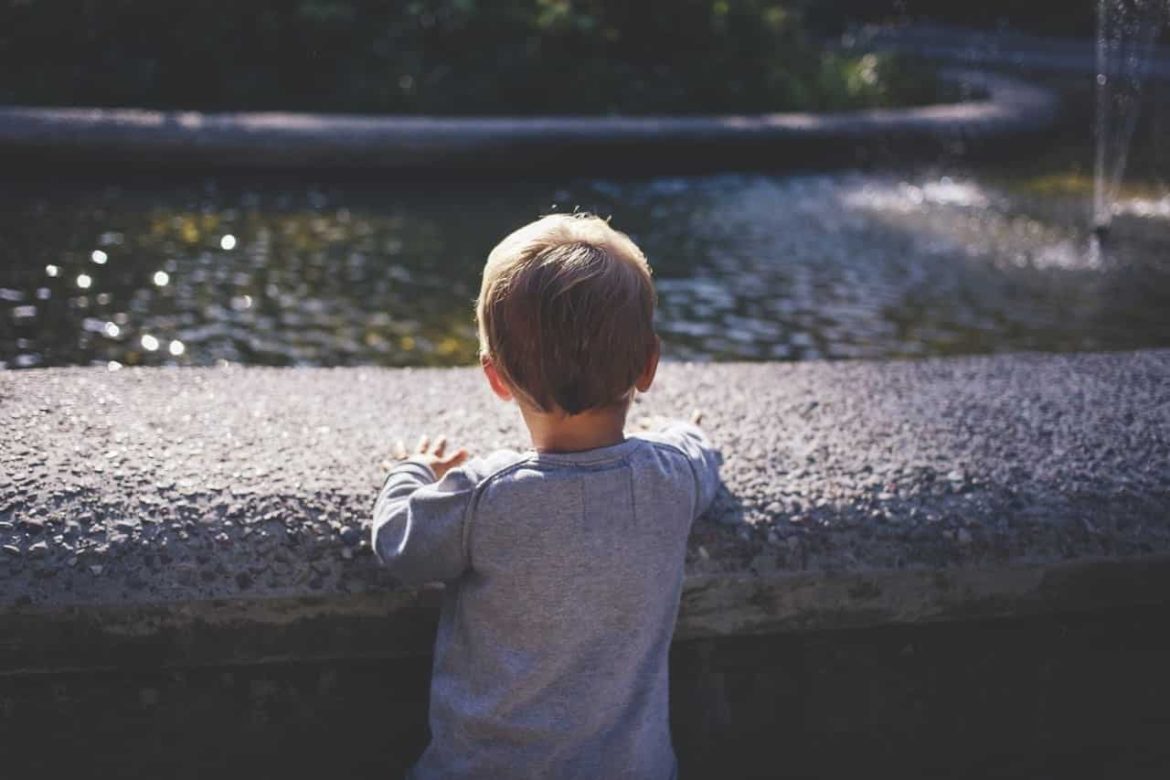 A young child with short blond hair and a gray shirt stands facing a stone edge, looking at a pond or fountain in a park, with sunlight casting shadows.