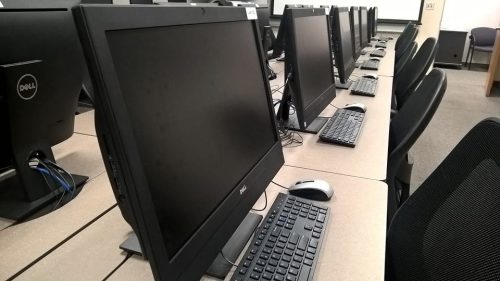 A row of desktop computers with monitors, keyboards, and mice are set up on desks in a computer lab. Empty chairs are positioned at each workstation. The room appears clean and organized.