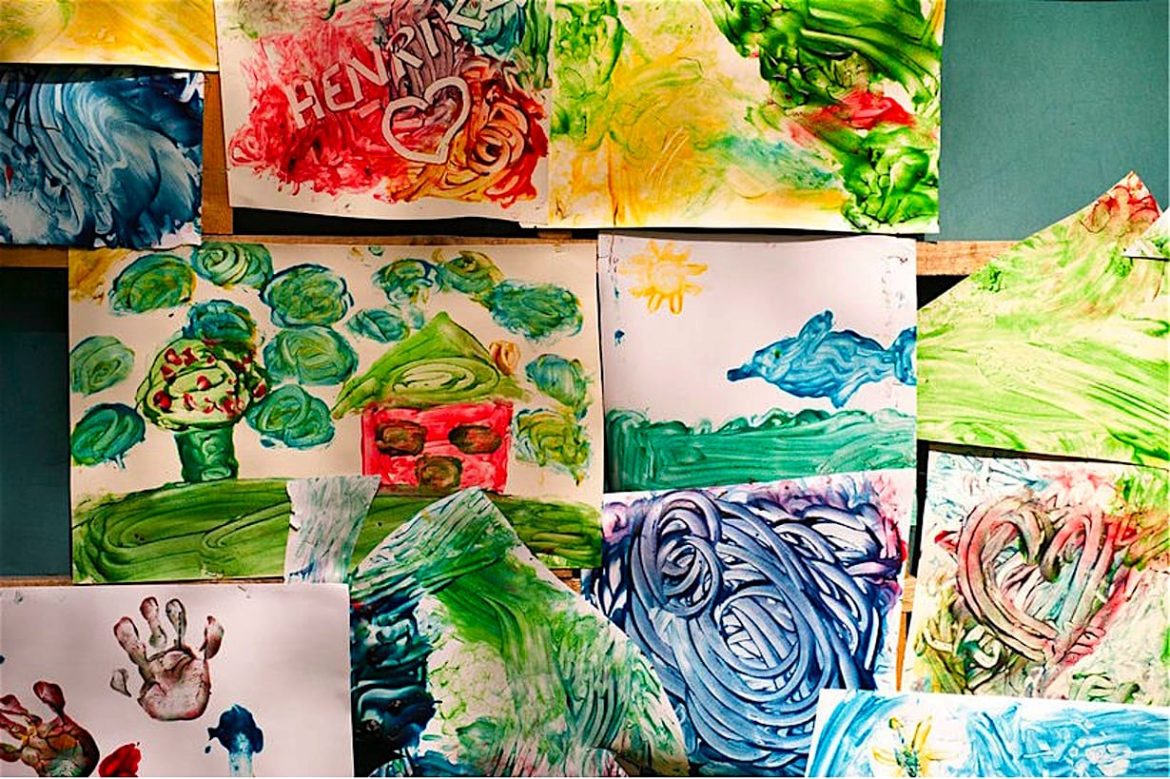 Several colorful childrens paintings are displayed on a wall, featuring handprints, hearts, a house with trees, swirling patterns, and a dolphin under a sunny sky. The artwork uses vibrant greens, blues, reds, and yellows.