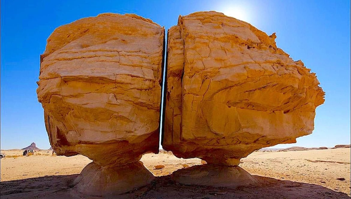 A large rock formation in a desert, split almost perfectly down the middle into two halves, both balanced on stone bases, with clear blue sky and sunlight in the background.