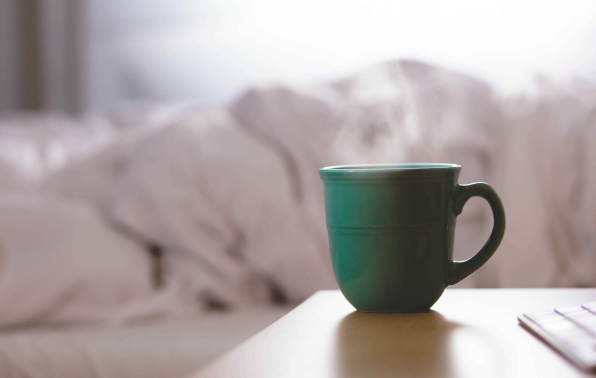 A green ceramic mug sits on a light wooden table in front of a bed with white, rumpled blankets in a softly lit, cozy room.