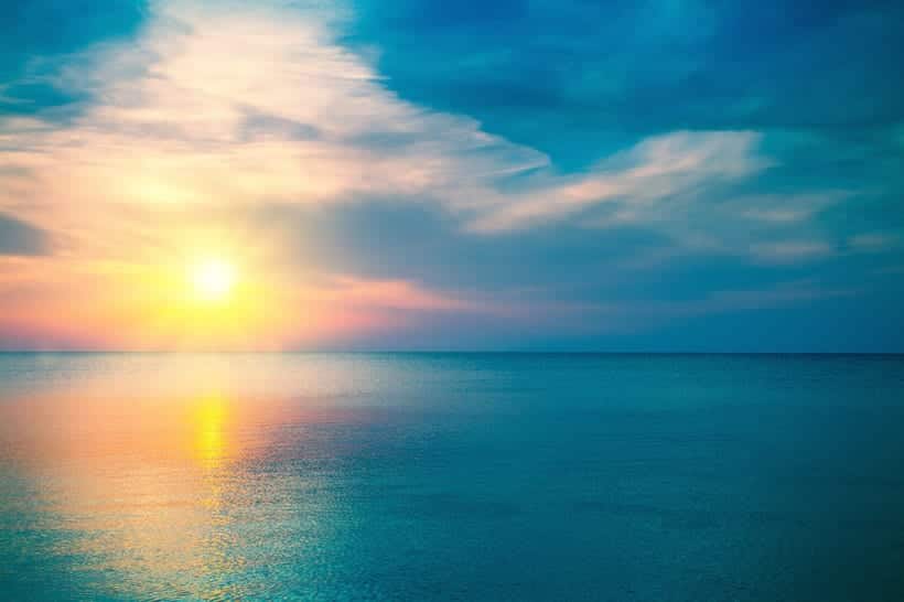 A vibrant sunset over a calm ocean, with the sun low on the horizon and colorful clouds creating a gradient of blue, yellow, pink, and orange in the sky, reflected softly on the water’s surface.