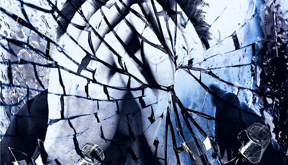 A black-and-white photo of a person is seen through a shattered glass pane, creating a fractured, fragmented effect. The image has a blue tint, with water drops and reflections on the broken glass pieces.