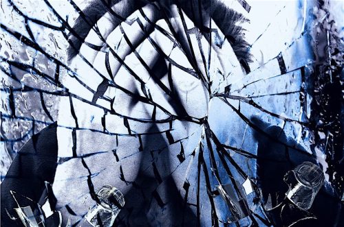 A black-and-white photo of a person is seen through a shattered glass pane, creating a fractured, fragmented effect. The image has a blue tint, with water drops and reflections on the broken glass pieces.