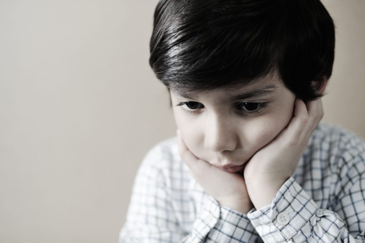 A young boy with dark hair, wearing a light plaid shirt, rests his chin on his hands and looks downward with a thoughtful, sad expression against a plain background.