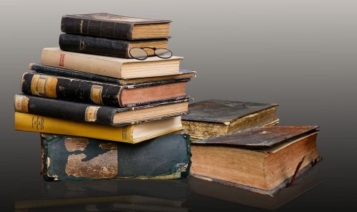 A stack of old, worn books sits on a surface, with a pair of round eyeglasses resting on top. Two additional aged books lie beside the stack, all against a neutral background.