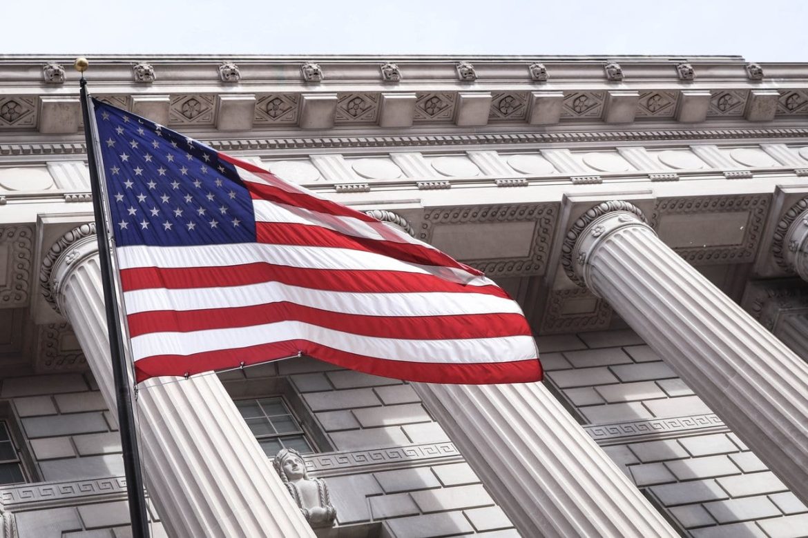 The American flag waves in front of a large, classical-style building with tall columns and ornate architectural details.