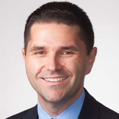 A man with short dark hair, wearing a dark suit, light blue shirt, and no tie, is smiling at the camera against a plain, light background.