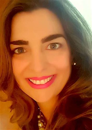 A woman with long brown hair smiles at the camera. She is wearing bright pink lipstick, has well-defined eyebrows, and is accessorized with a pearl necklace and pearl earrings. The background is softly lit.