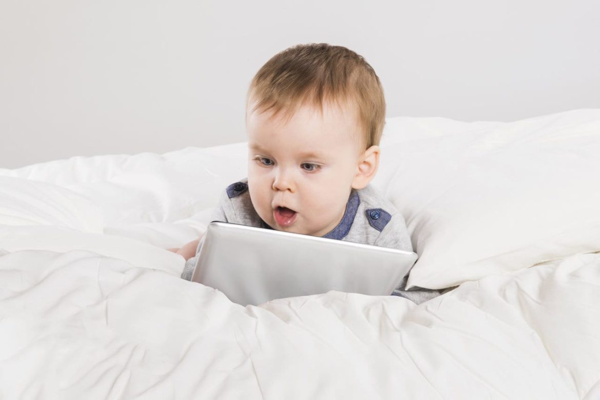 A baby lies on a white bed, looking intently at a tablet with an open-mouthed expression of surprise or curiosity.