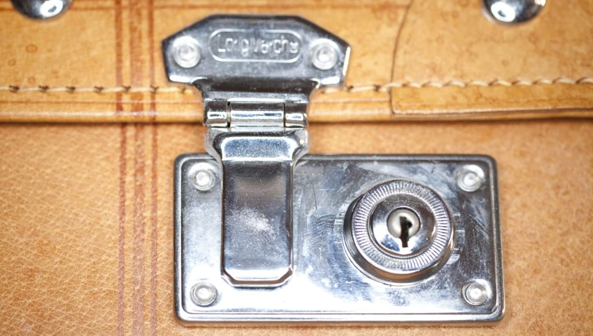 Close-up of a shiny silver latch and keyhole lock on a light brown leather suitcase or briefcase, showing stitching detail and metal hardware.