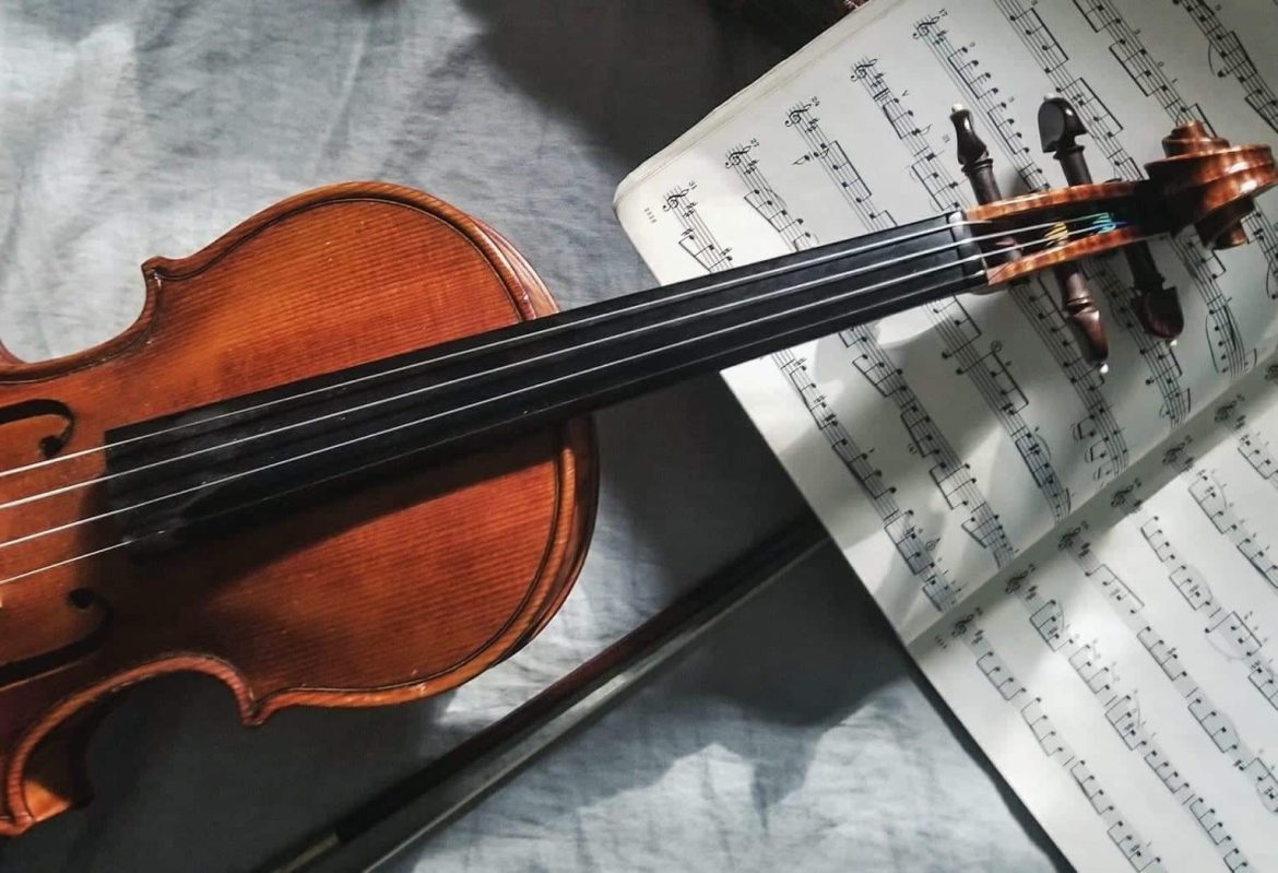 A wooden violin lies on a gray surface next to an open sheet music book, with a bow resting underneath the instrument. Sunlight partially illuminates the scene.