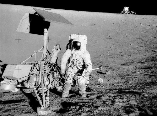 An astronaut in a spacesuit stands next to a robotic lunar probe on the Moon’s surface. The ground is dusty, and another spacecraft is visible in the background on the lunar horizon.