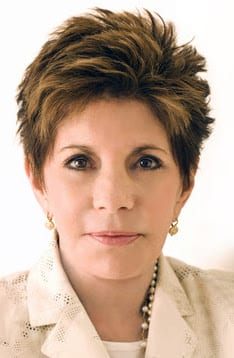 A woman with short, brown, styled hair is wearing a cream-colored jacket, pearl earrings, and a pearl necklace. She is facing forward against a plain, light background.