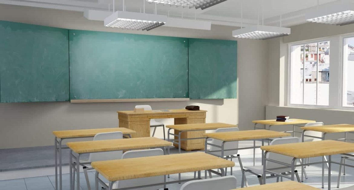 Empty classroom with wooden desks and white chairs facing a large green chalkboard, a wooden teacher’s desk, and windows letting in natural light. The room has white walls, bright ceiling lights, and a clean, organized appearance.
