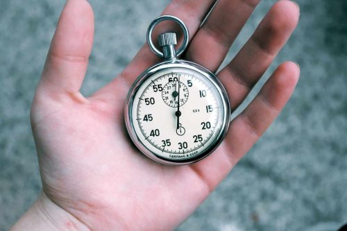 A hand holding a silver analog stopwatch with a white face, black numbers, and a winding crown, against a blurred gray background.