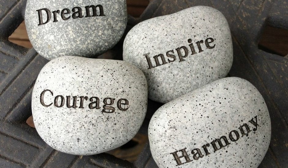 Four smooth gray stones with engraved words: Dream, Inspire, Courage, and Harmony, arranged closely together on a textured dark surface.