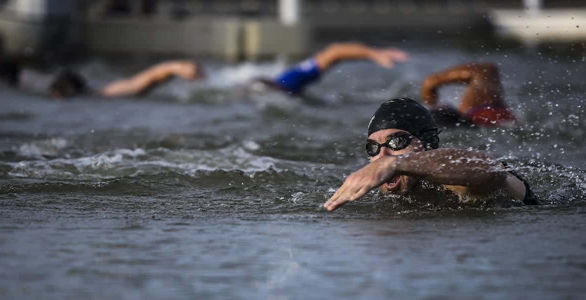 Multiple swimmers wearing swim caps and goggles compete in an open water race, swimming front crawl in choppy water near a dock or pier. The focus is on the swimmer in the foreground.