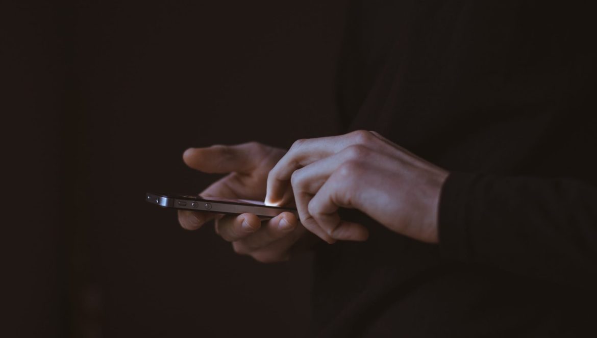 A person wearing a dark long-sleeve shirt is using a smartphone, touching the screen with one hand, against a dark background. Only their hands and part of their torso are visible.