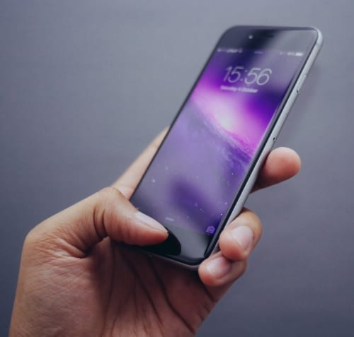 A hand holds a smartphone with a purple galaxy wallpaper. The screen displays the time as 15:56 and the date as Sunday, 9 October. The background is plain and gray.