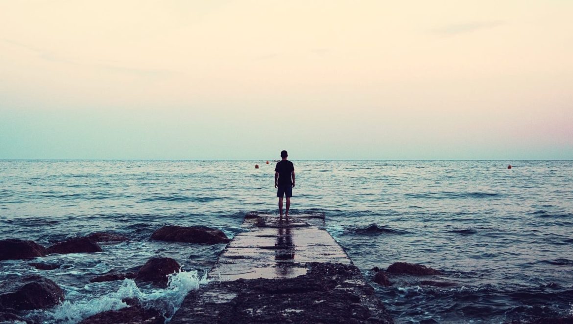 A person stands at the end of a rocky pier facing the calm sea under a pastel-colored sky at sunset, with gentle waves surrounding the rocks.