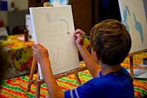 A child in a blue shirt sits at a table and paints the outline of a cat on a canvas set on an easel. The table is covered with a colorful, patterned cloth.