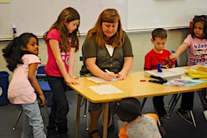 A teacher sits at a table in a classroom, writing on paper, while four young children stand and sit around her, watching and participating in an activity.