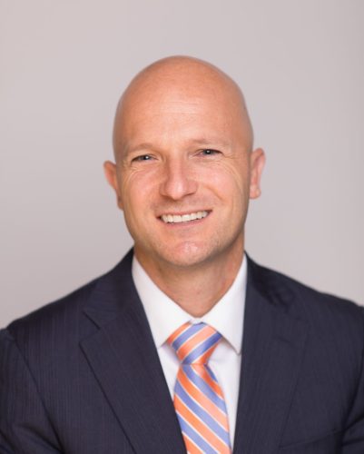 A bald man in a dark suit and orange-striped tie smiles at the camera against a plain light-colored background.
