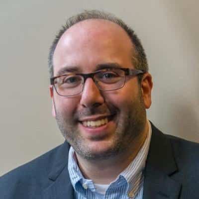 A man with glasses, short brown hair, and a beard, wearing a dark suit jacket, a blue striped shirt, and smiling in front of a neutral background.