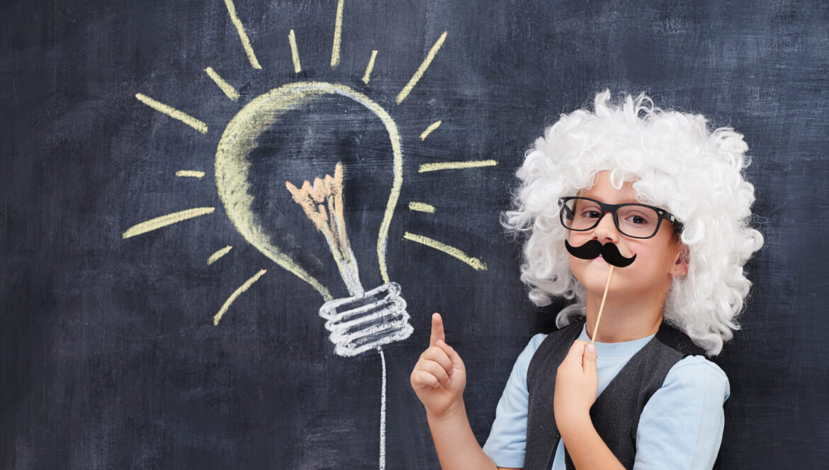 A child wearing a white curly wig, black glasses, and a fake mustache stands in front of a chalkboard with a large, glowing lightbulb drawn on it, pointing upward as if having an idea.