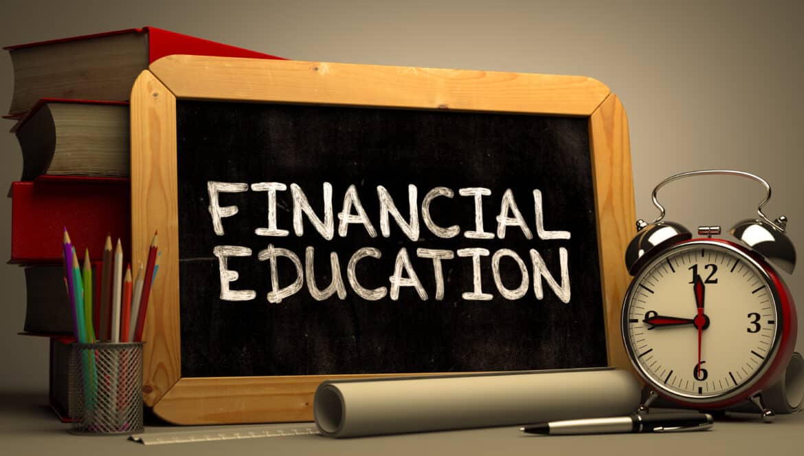 A chalkboard with Financial Education written on it sits on a table next to stacked books, colored pencils in a holder, a rolled-up paper, and a classic alarm clock showing 7:00.
