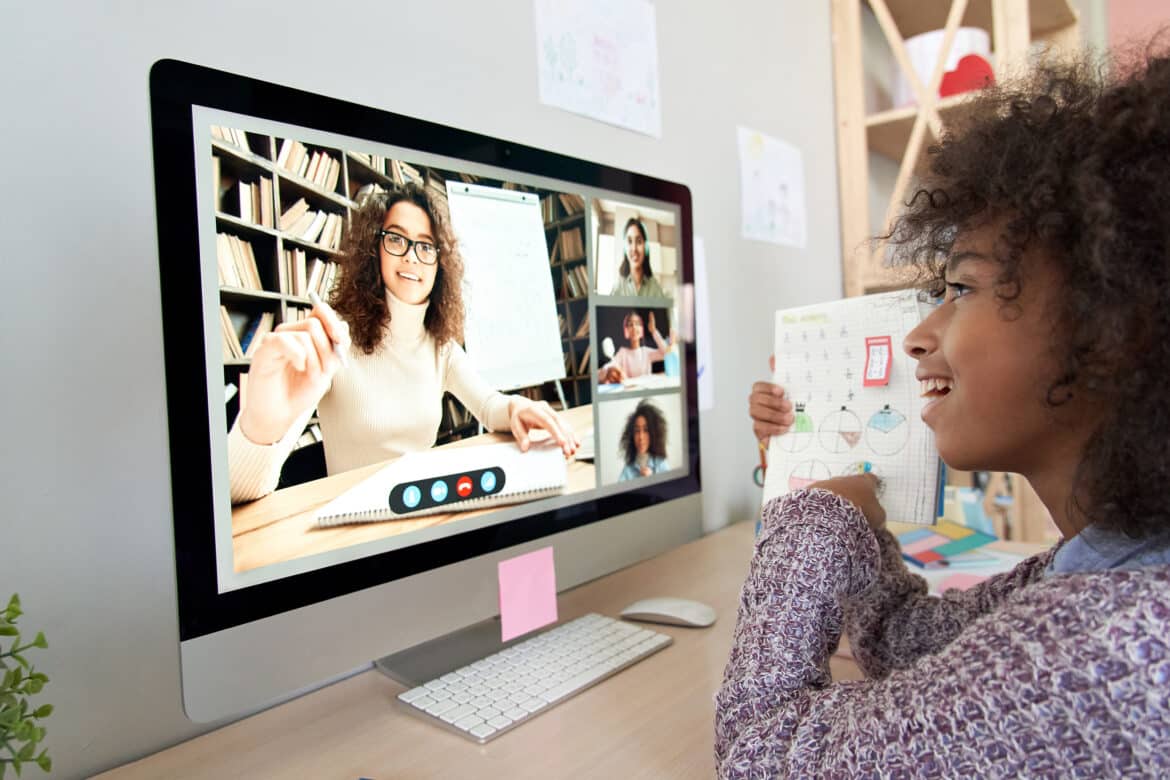 A girl holds up a math worksheet while participating in an online class on her computer, interacting with a teacher and classmates visible on the monitor during a video call.