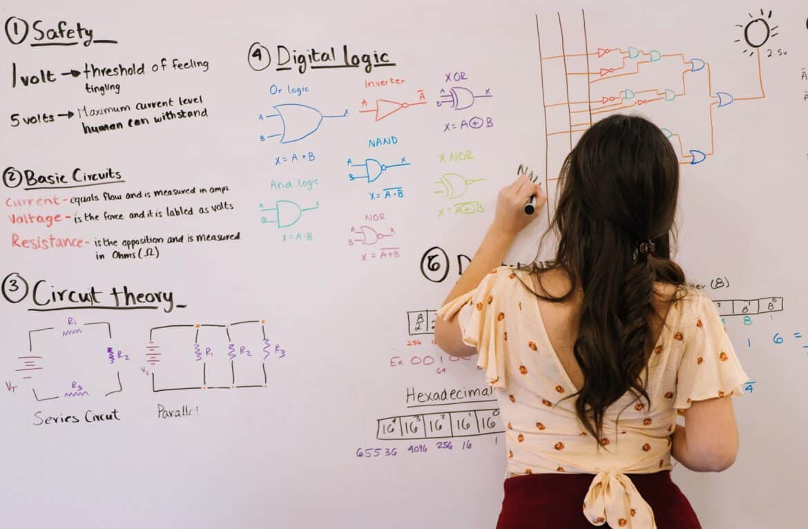 A woman with long brown hair writes on a whiteboard covered with colorful diagrams and notes about safety, basic circuits, circuit theory, digital logic, and hexadecimal numbers.