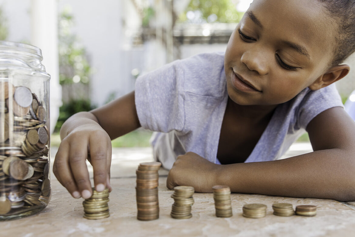 A child lies on the ground, smiling while stacking and organizing coins into piles. A large glass jar filled with coins sits beside them. Sunlight filters through trees in the background.