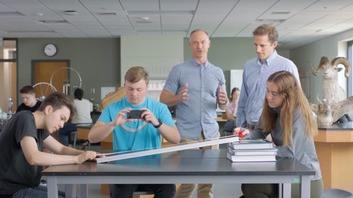 Four students conduct a physics experiment with a ramp and books on a table, while two adults observe and guide them in a classroom filled with scientific equipment and animal models.