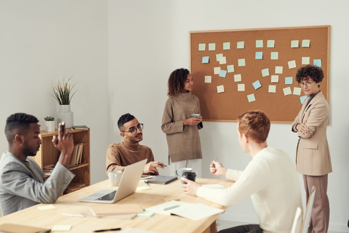Five people are having a discussion in an office. Four sit at a table with laptops and notebooks, while one stands by a corkboard covered with sticky notes, engaging the group.