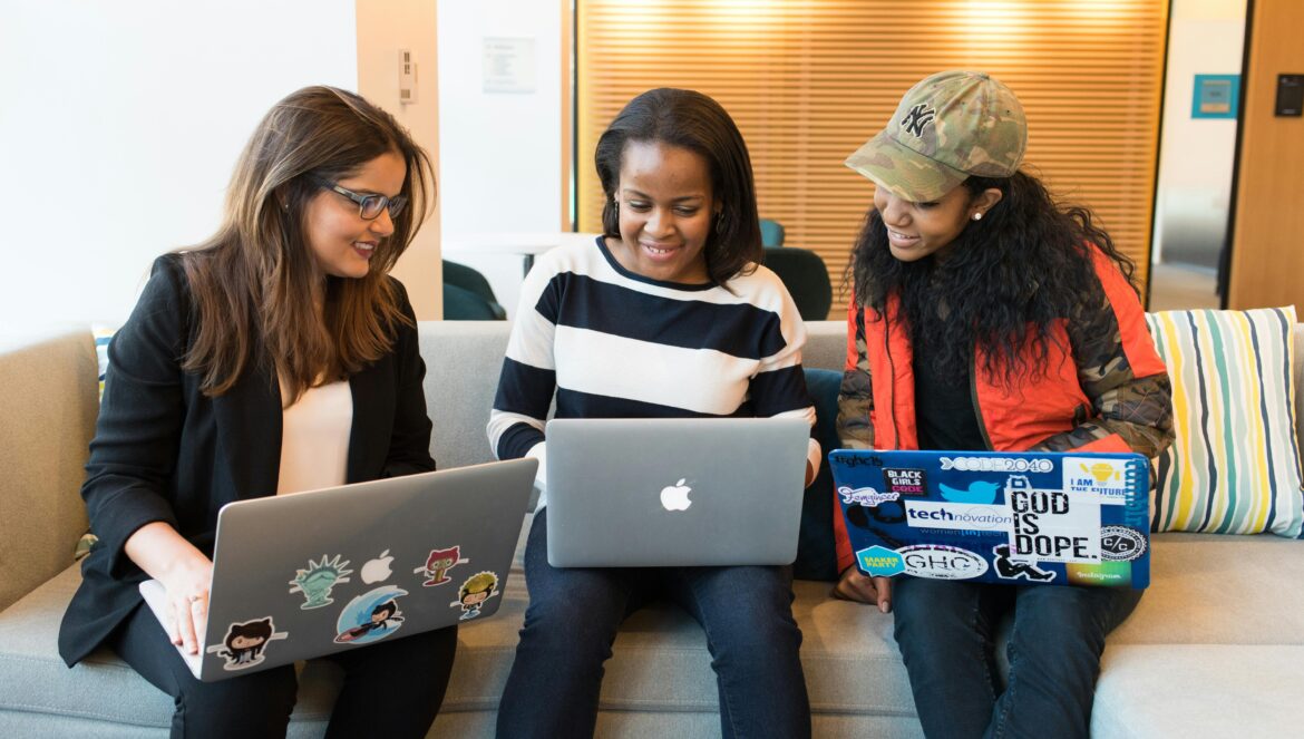 Three women sit on a couch using laptops, smiling and collaborating. Two laptops are decorated with stickers, and the background shows a modern office with wooden blinds and colorful cushions.