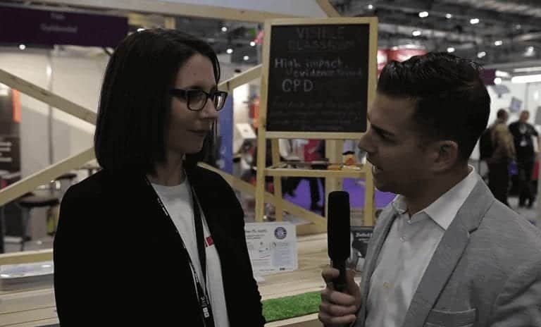 A woman and a man are having an interview or conversation at an indoor event. The man holds a microphone and faces the woman, who wears glasses. An exhibition booth and a chalkboard sign are visible in the background.