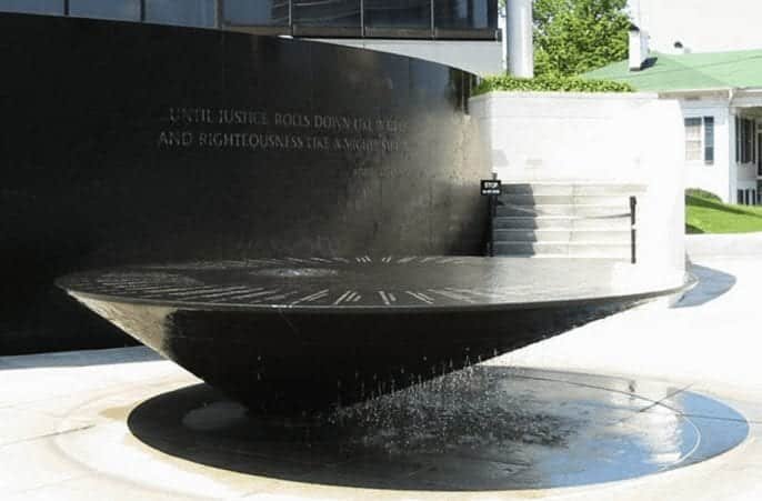 A circular black granite fountain with water flowing over its edge set in front of a curved black stone wall with engraved text Steps and green trees are visible in the background