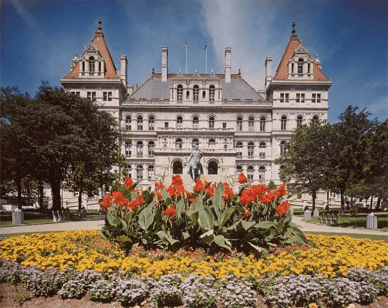 A large historic building with towers and ornate details stands behind a colorful flower garden featuring red, yellow, and purple blooms, surrounded by trees under a blue sky.