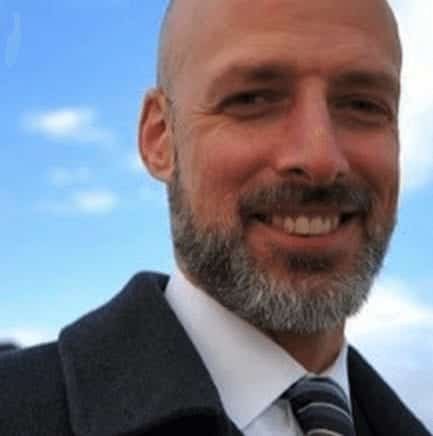 A smiling man with a bald head and beard, dressed in a suit and tie, stands outdoors against a blue sky with scattered clouds.
