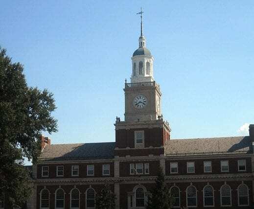A historic red-brick building with white trim and a prominent clock tower topped with a dome, set against a clear blue sky, with trees partially visible in the foreground.