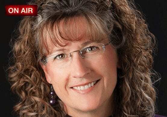 A woman with curly brown hair, glasses, and a friendly smile is shown against a dark background. She is wearing a purple top and earrings. A red “ON AIR” sign appears in the top left corner.