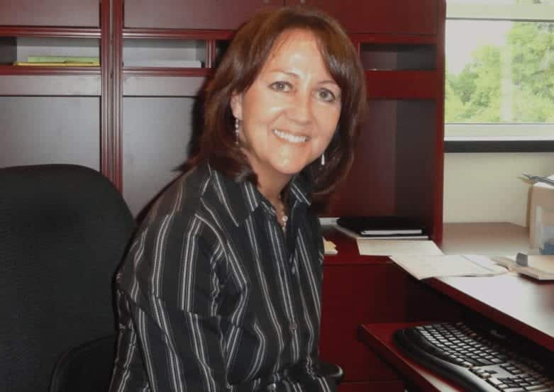 A woman with brown hair, wearing a black and white striped shirt, sits and smiles at a desk with a computer keyboard and office supplies. There is a window with greenery visible in the background.