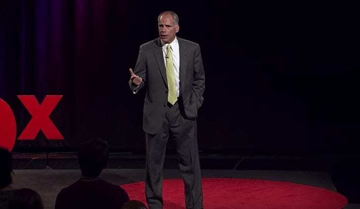 A man in a gray suit and yellow tie stands on a red circular carpet, speaking and gesturing with one hand on a stage with a dark background and a partial red TEDx logo visible.